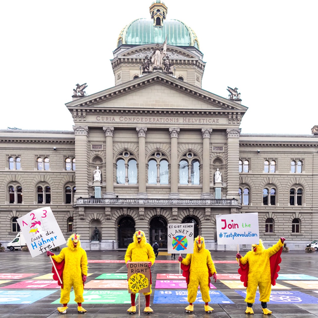 Chickens in costume holding signs including 'Join the #TastyRevolution' outside the Swiss Federal Palace